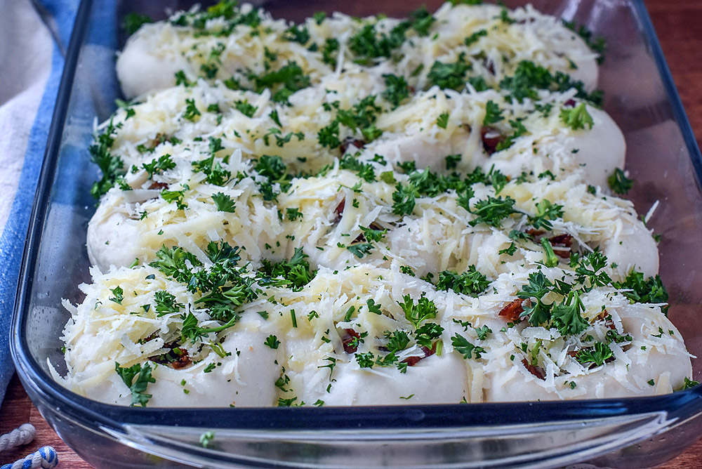 A pan of fresh dinner roll dough topped with cheese, bacon, and parsley.