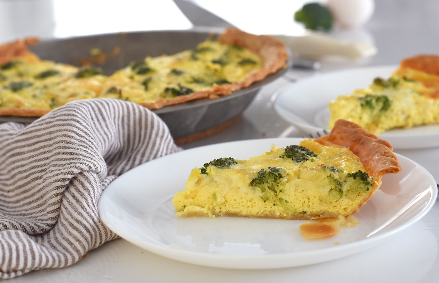 broccoli and cheese quiche slice on a plate with a pan of quiche in the background